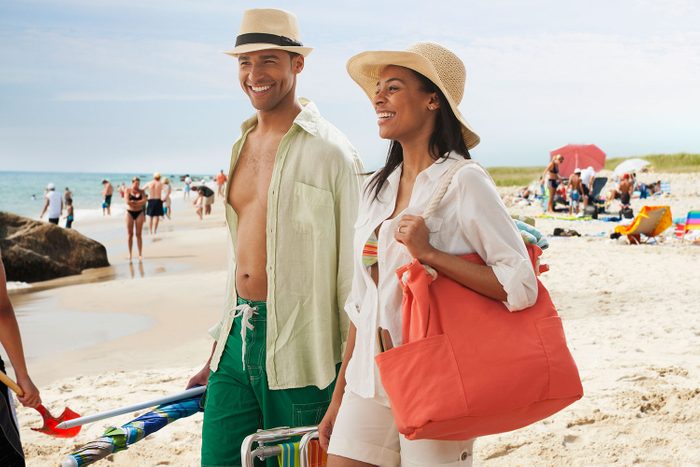 Couple Walking On Beach in summer outfits with beach umbrella, coole and beach bag in hand