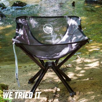 A portable folding chair sits on sandy ground beside a calm stream, with sunlight filtering through nearby trees, creating a serene outdoor atmosphere.