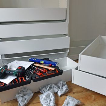 A white drawer unit sits partially assembled, with tools and assembly instructions scattered nearby on a wooden floor, indicating ongoing construction or setup.