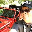 A person wearing sunglasses and a cap takes a selfie next to a red vehicle, surrounded by greenery.