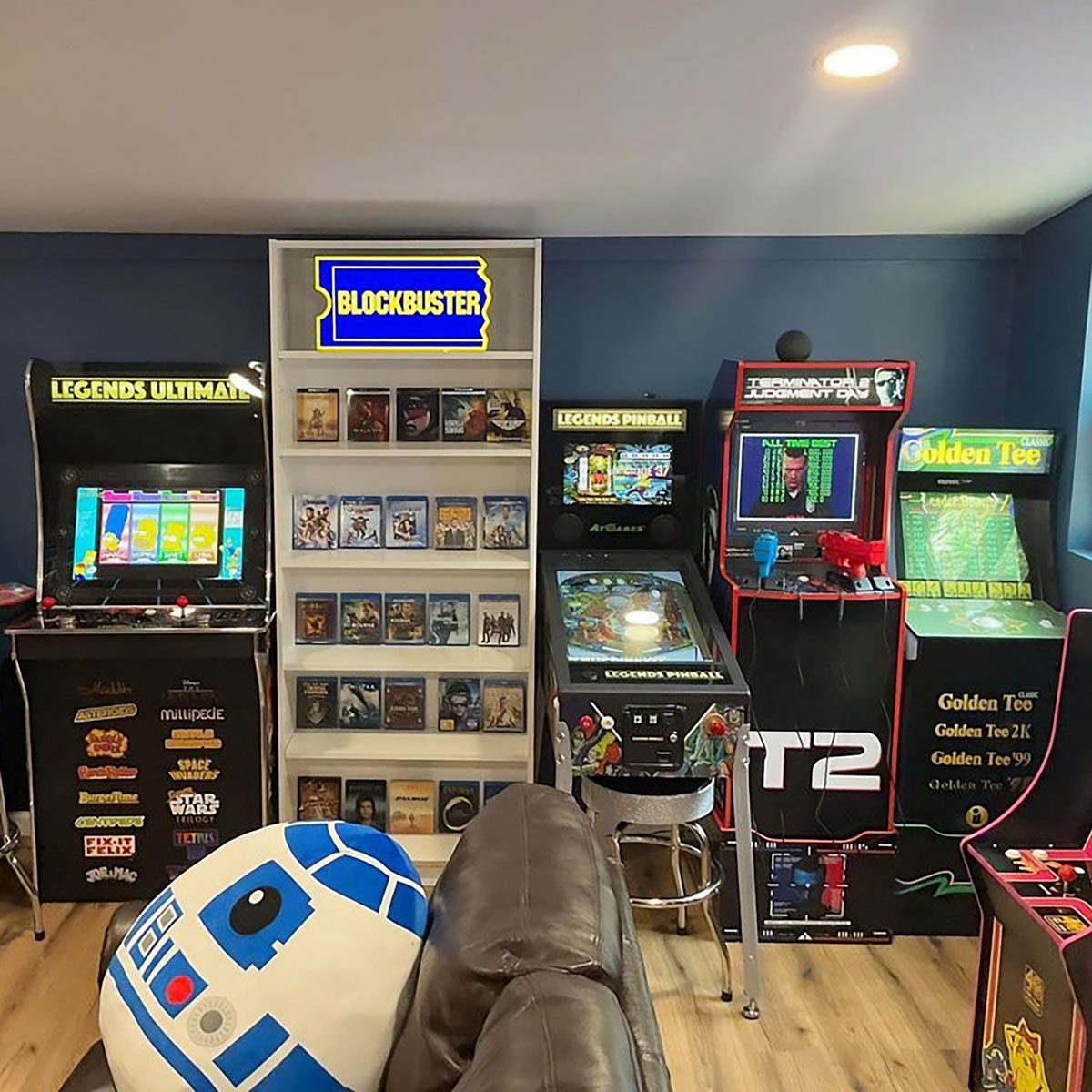 An arcade room features multiple gaming cabinets, a bookshelf of DVDs, and a decorative "Blockbuster" sign, creating a nostalgic entertainment environment.