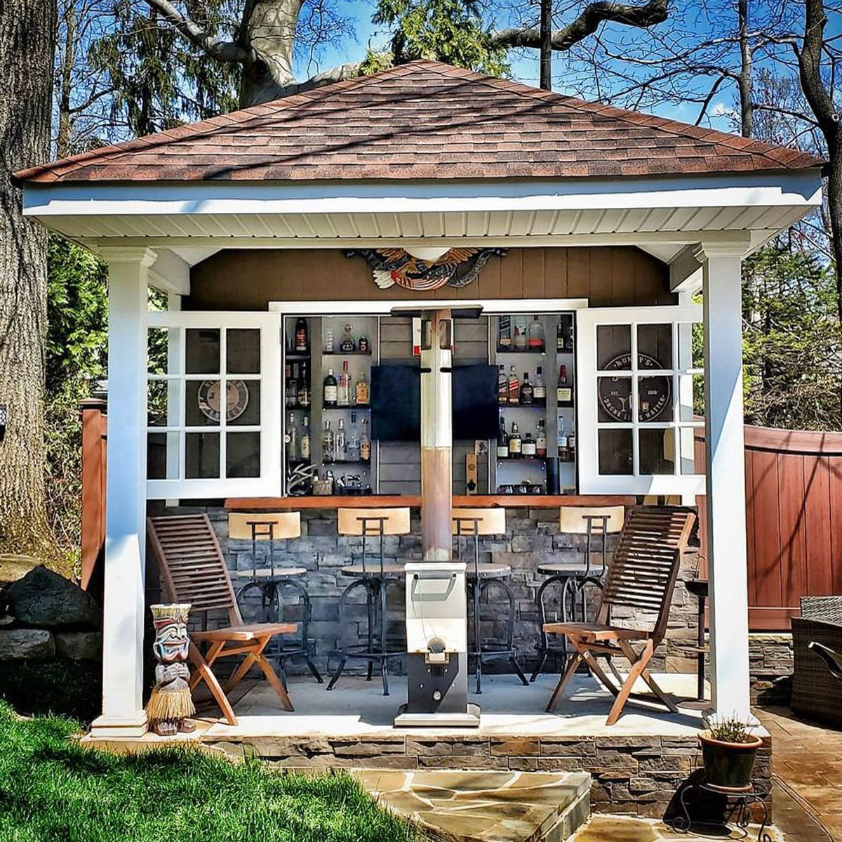 A gazebo hosts a bar with shelves of bottles, two stools, and comfortable seating, surrounded by lush greenery and a wooden fence.