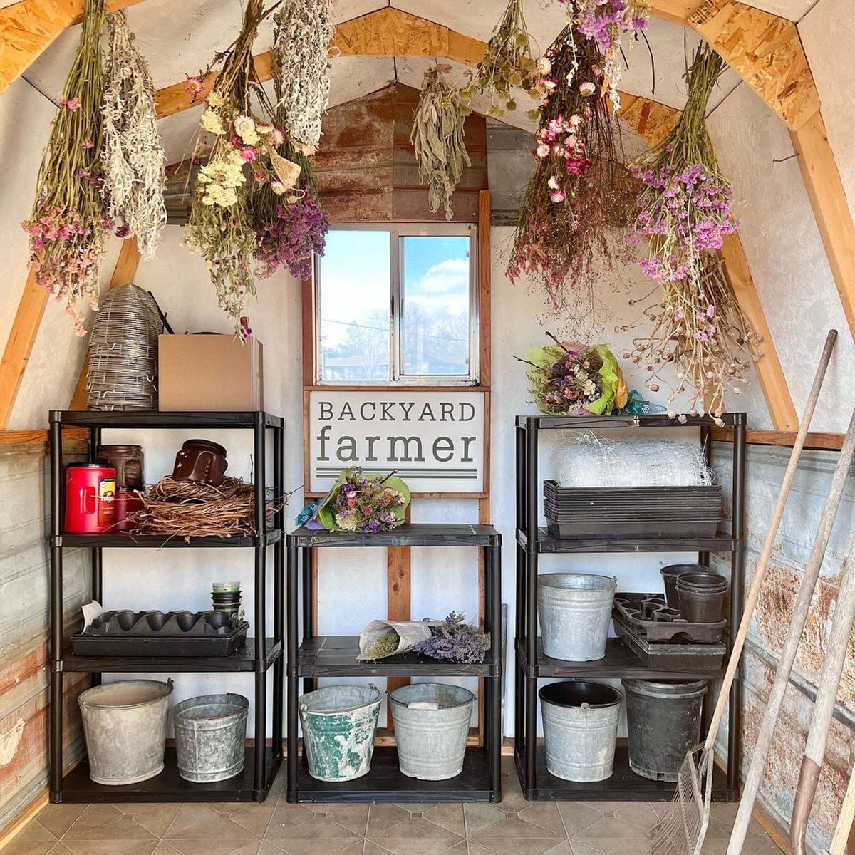 Dried flowers hang above metal shelves filled with gardening tools and containers in a rustic storage space, with a sign reading "BACKYARD farmer" on the wall.