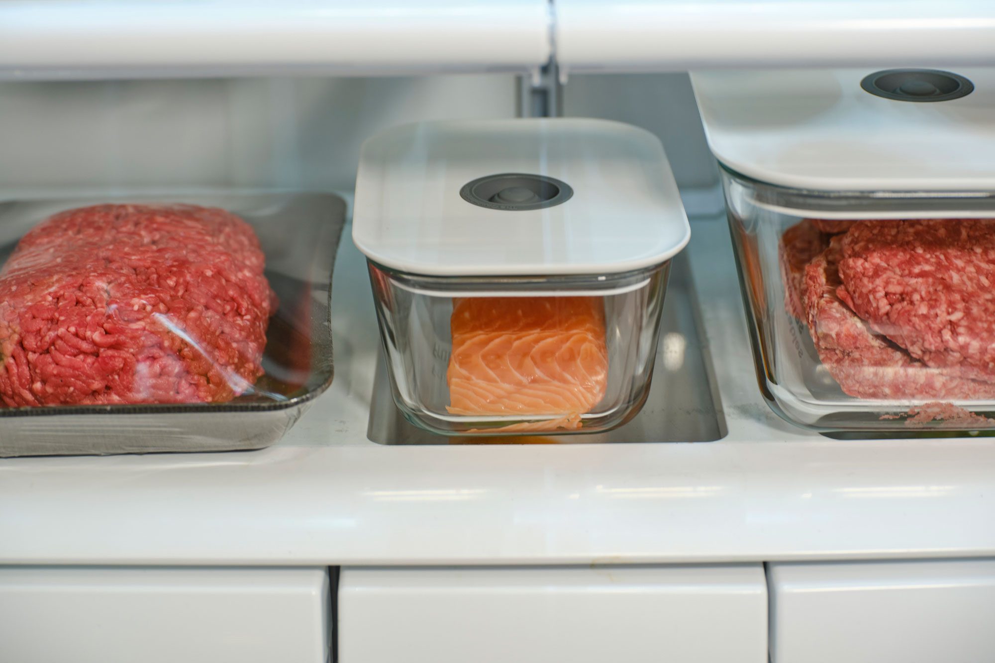 Raw meats, including ground beef and salmon, are stored in glass containers within a refrigerator, displaying organization and preservation of food items.