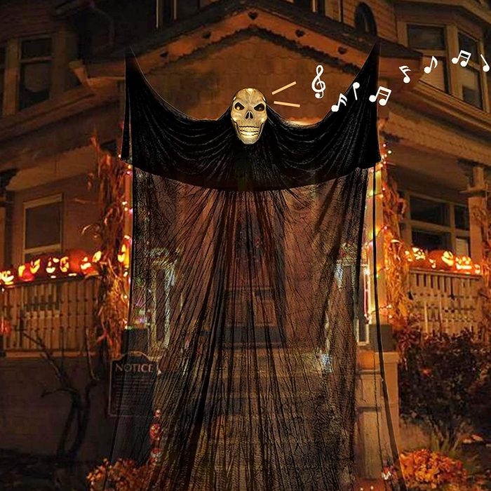 A black, ghost-like figure with a skull head sways while emitting music, positioned in a festive, decorated outdoor setting with pumpkins and lights.