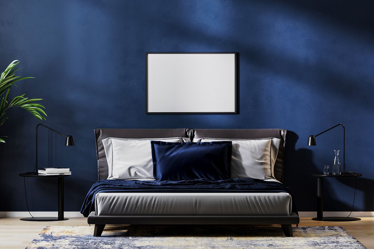 A bed with dark bedding rests against a navy wall, flanked by side tables and reading lamps, with a decorative plant nearby.