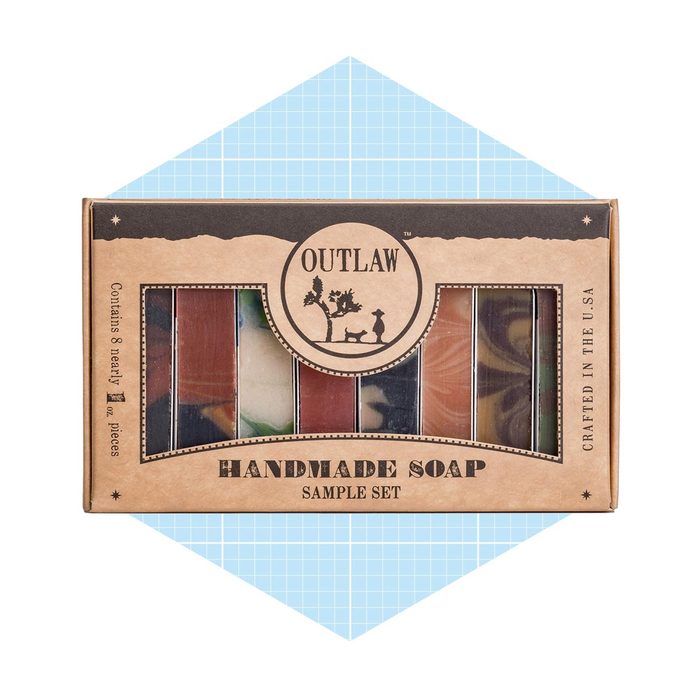 A boxed set of handmade soap bars is displayed, showcasing eight colorful pieces, against a light blue geometric background.