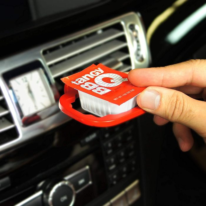 A hand holds a red condiment container while placing it near a car's dashboard, which features a clock and audio controls in a dark interior.