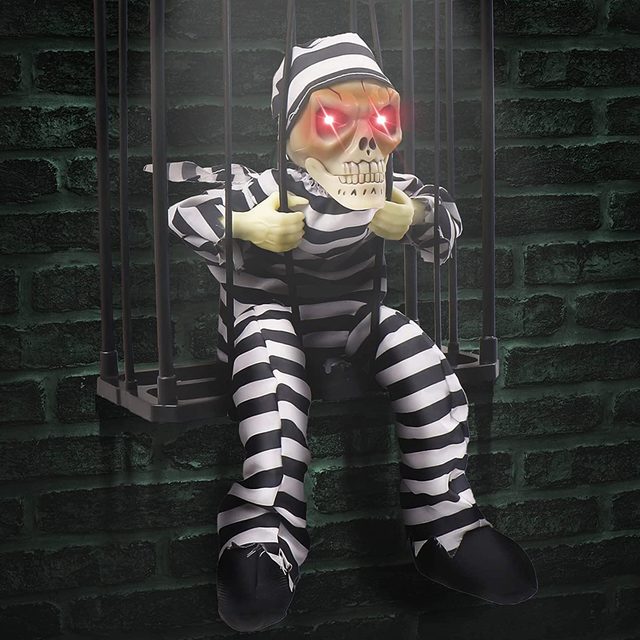 A skeleton figure in striped prison attire sits in a barred cage, gripping the bars with glowing red eyes, against a dark brick wall backdrop.