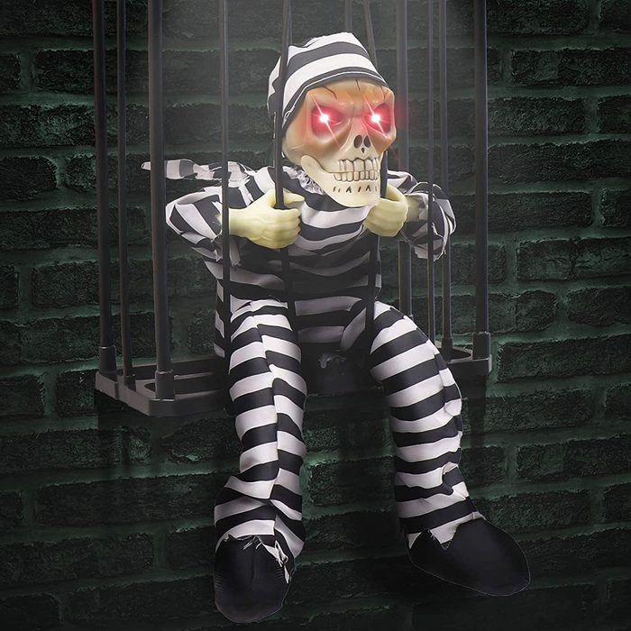 A skeleton figure in striped prison attire sits in a barred cage, gripping the bars with glowing red eyes, against a dark brick wall backdrop.