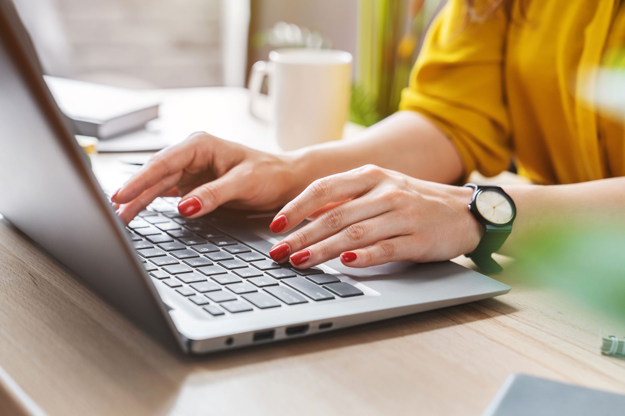 A person types on a laptop, wearing a watch and displaying red nails, while seated at a wooden desk with a cup and books nearby.