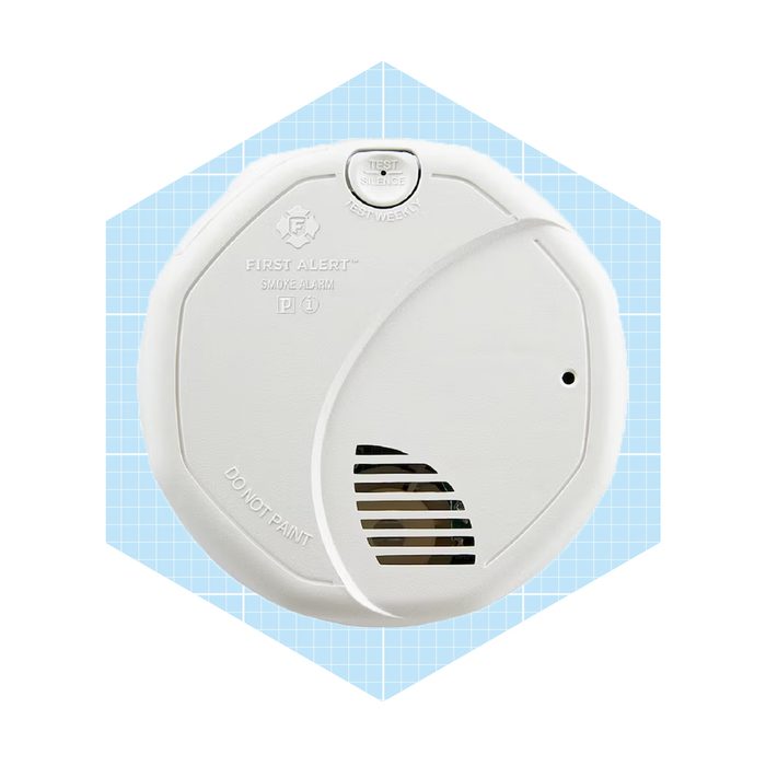 A white smoke alarm is mounted on a light blue grid-patterned background, featuring buttons and a vent for smoke detection.