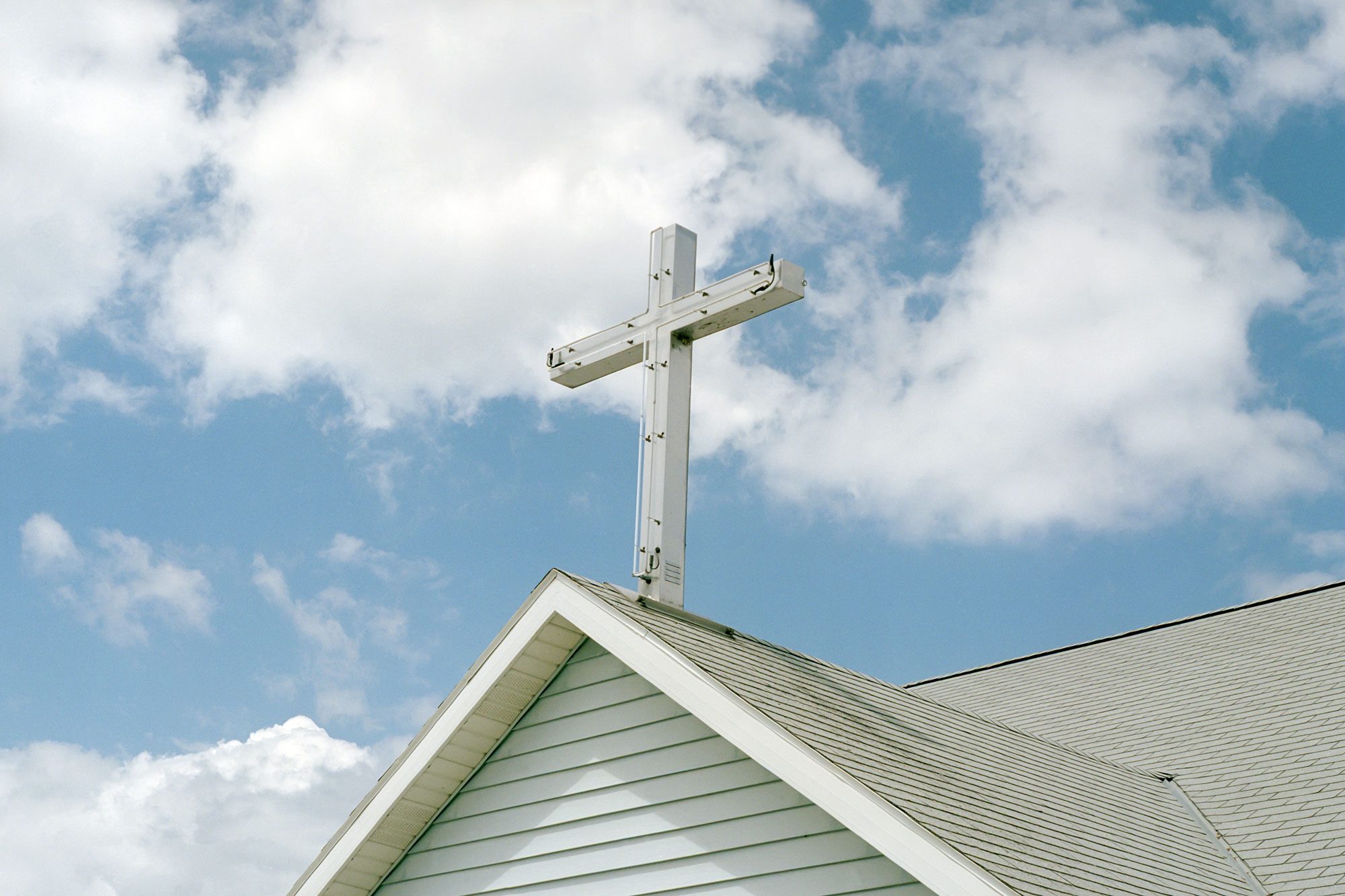 A white cross stands atop a pitched-roof church, reaching towards a blue sky filled with scattered clouds.