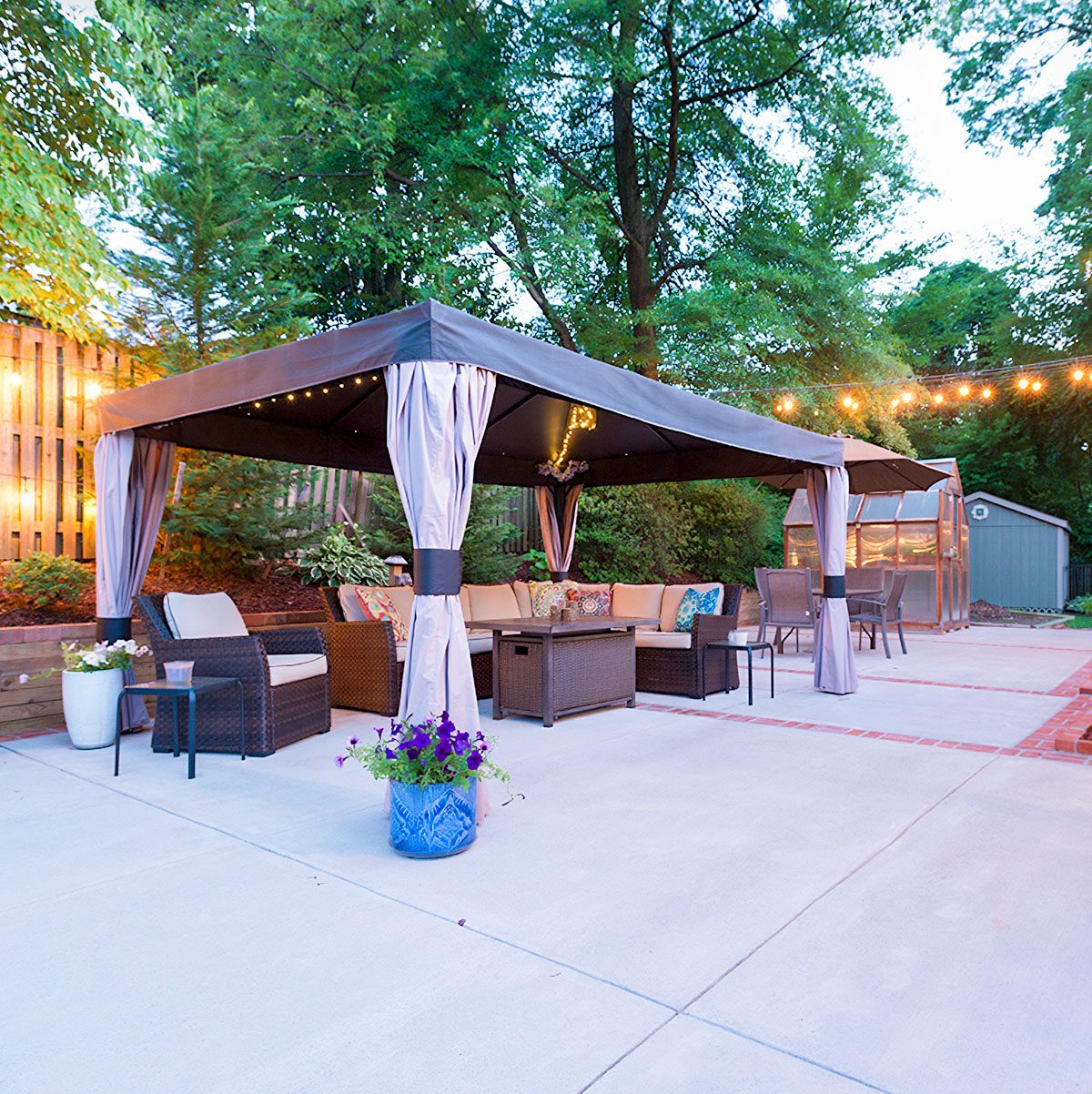 A gazebo with cushions and lights stands on a patio surrounded by greenery and a small shed, offering a cozy outdoor space for relaxation.