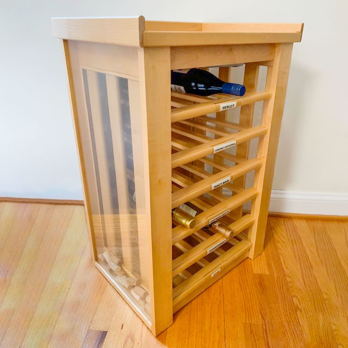 A wooden wine rack holds several bottles, with a glass front for visibility, placed on a hardwood floor against a light wall.