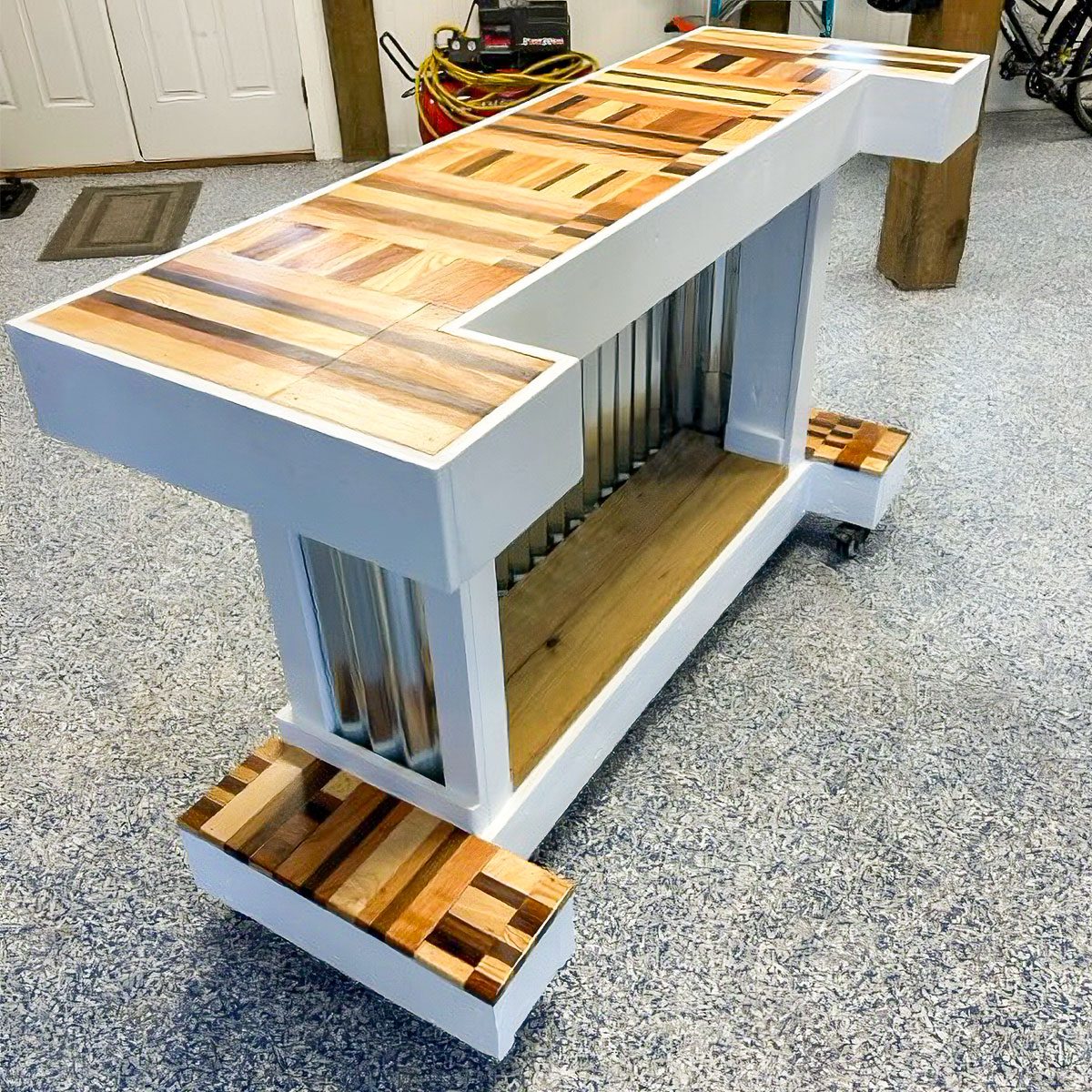 A wooden bar cart with a patterned top and metallic sides, on wheels in a garage setting, is ready for storage or serving purposes.