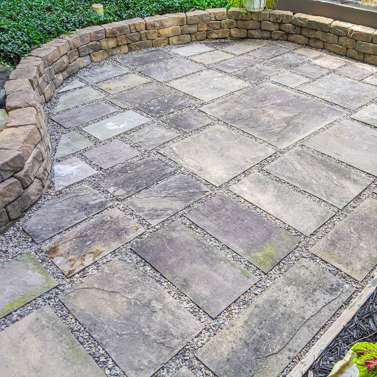 Paved stone patio with rectangular slabs and gravel spaces, surrounded by a low stone wall and greenery, offering a calm and structured outdoor space.