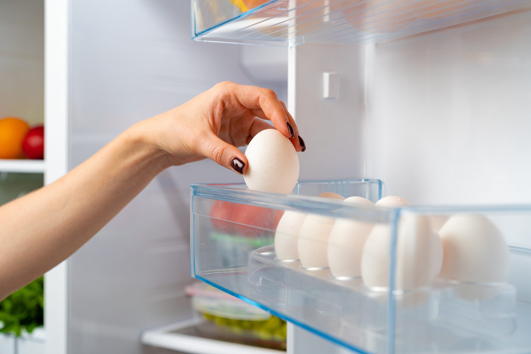 A hand lifts an egg from a clear refrigerator compartment, where several eggs are stored, amidst a background of various fruits and vegetables.