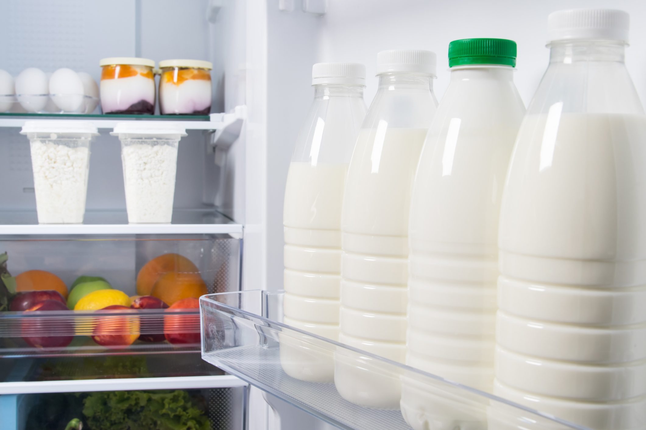Bottles of milk stand upright in a refrigerator, next to containers of food and fresh fruits, indicating a well-stocked interior.