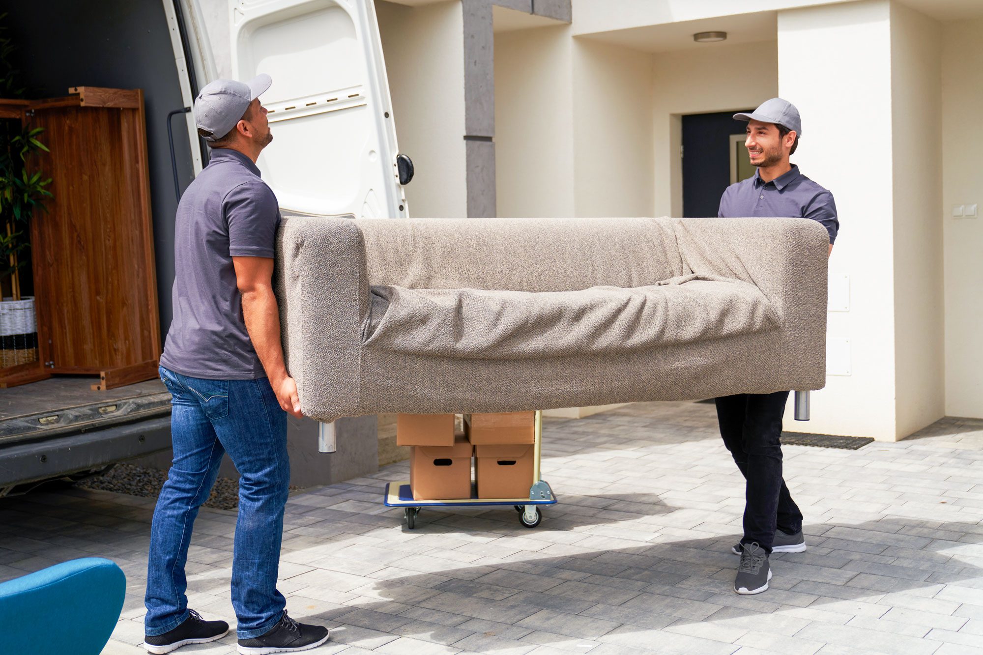 Two men lift a large sofa, carrying it from a delivery truck to a residential entrance, surrounded by a stone pathway and cardboard boxes.