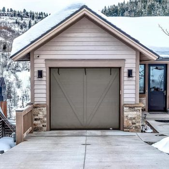 A garage door is closed, situated at the front of a house. Snow blankets the roof, while a driveway leads to the entrance amid a mountainous landscape.