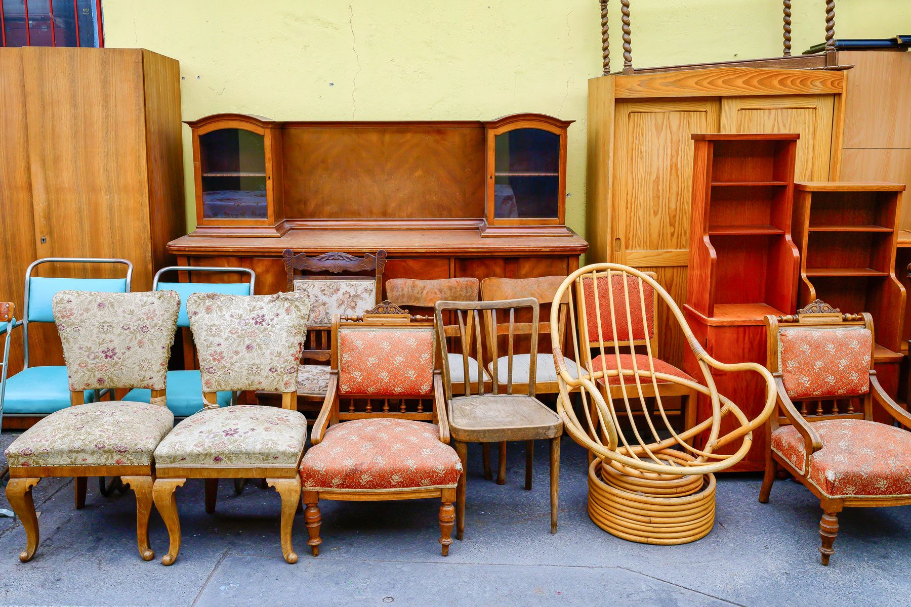 A variety of vintage chairs and furniture pieces are arranged against a yellow wall, showcasing diverse styles and materials in a cluttered setting.