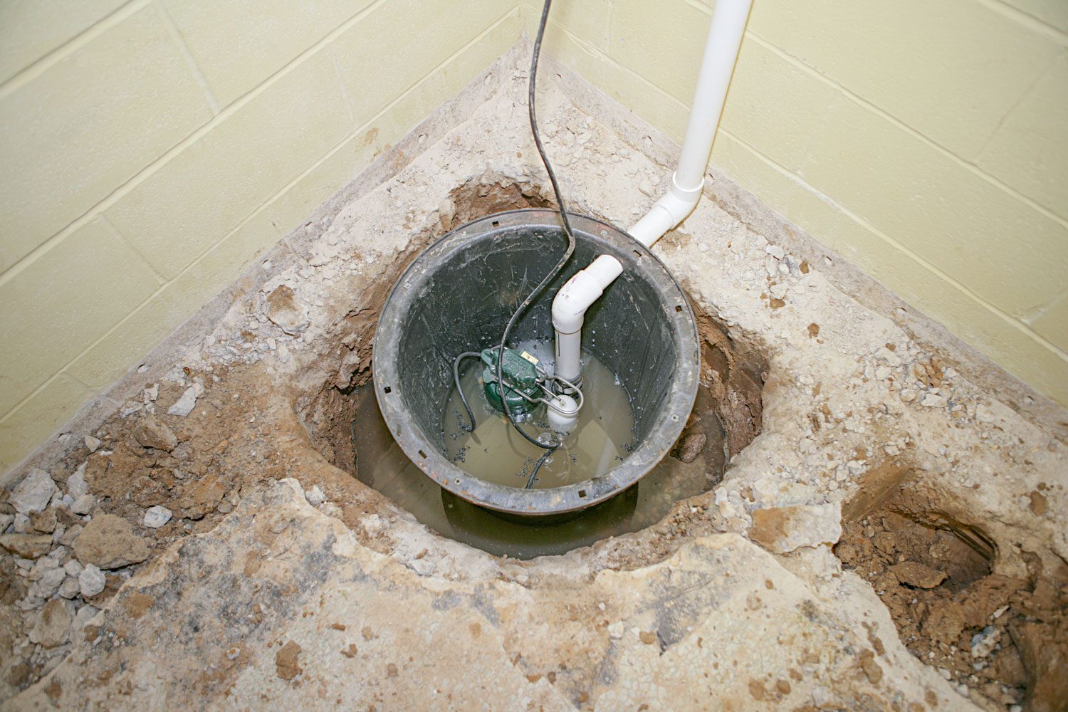 A sump pit filled with water contains a pump, surrounded by concrete debris, in a room with light green walls.