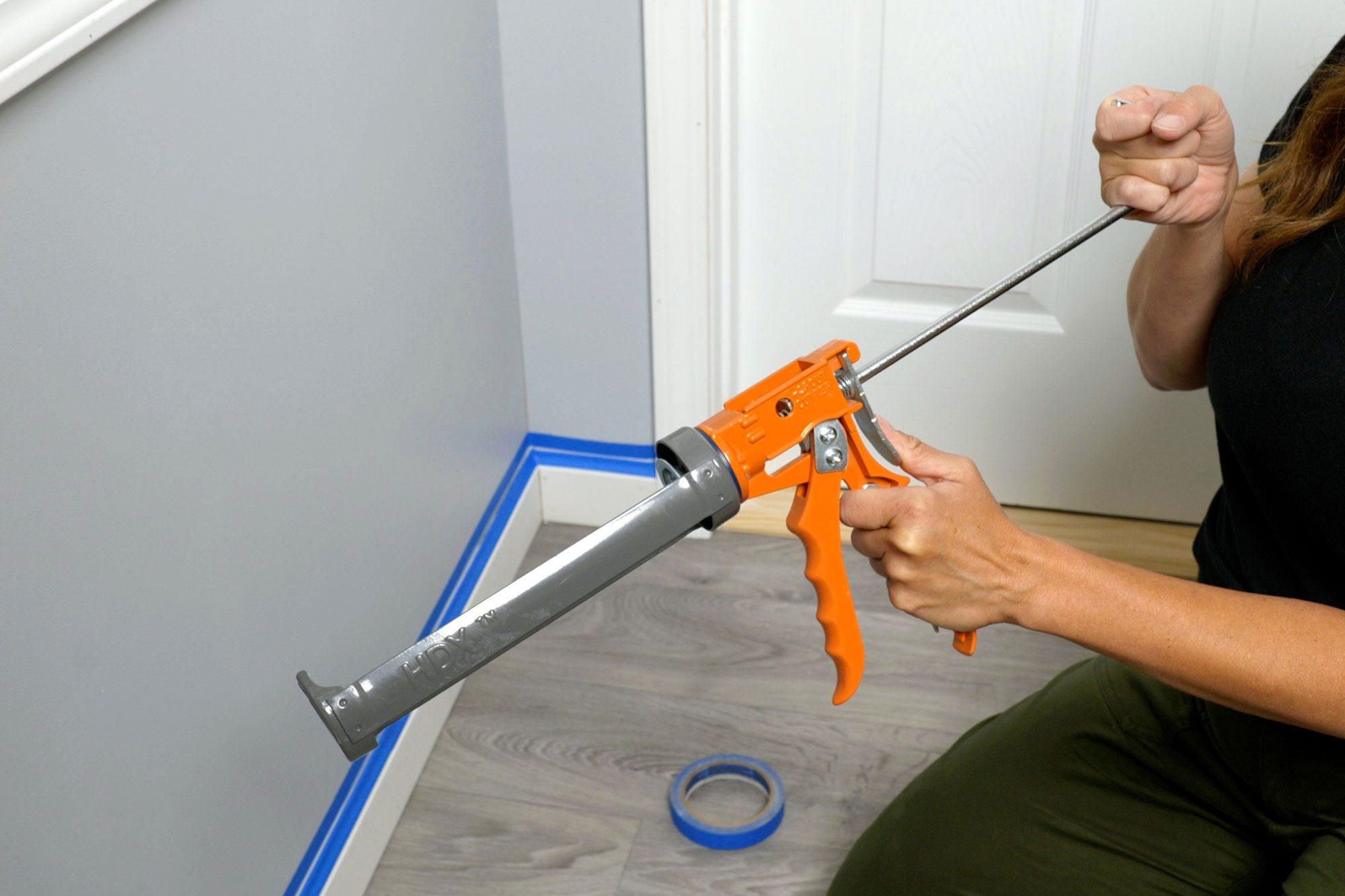 A person uses a caulking gun to apply sealant along a wall's baseboard in a room with gray walls and a light-colored floor.
