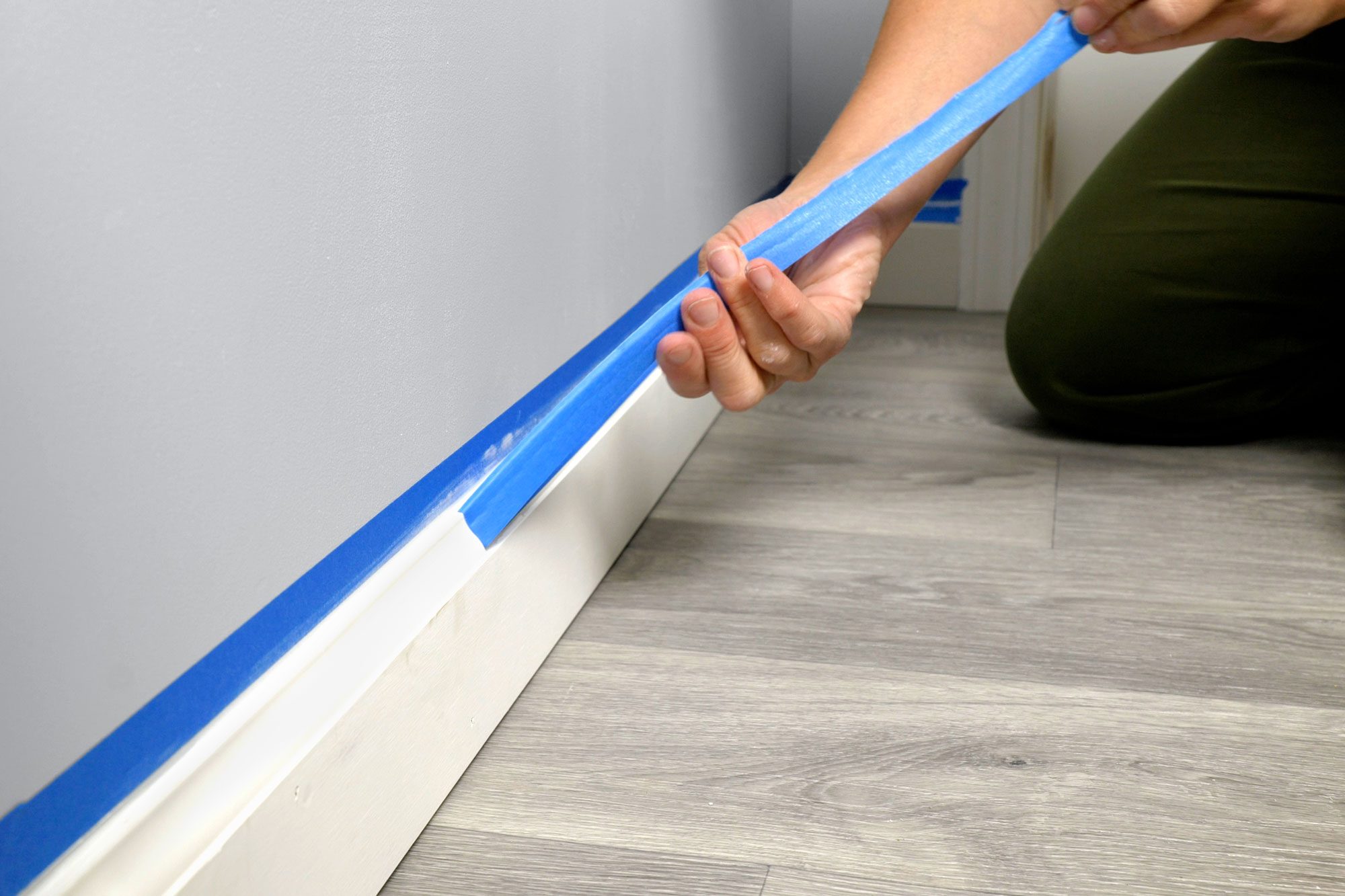 A hand applies blue painter's tape along a white baseboard against a gray wall, preparing for painting in a room with light wood flooring.
