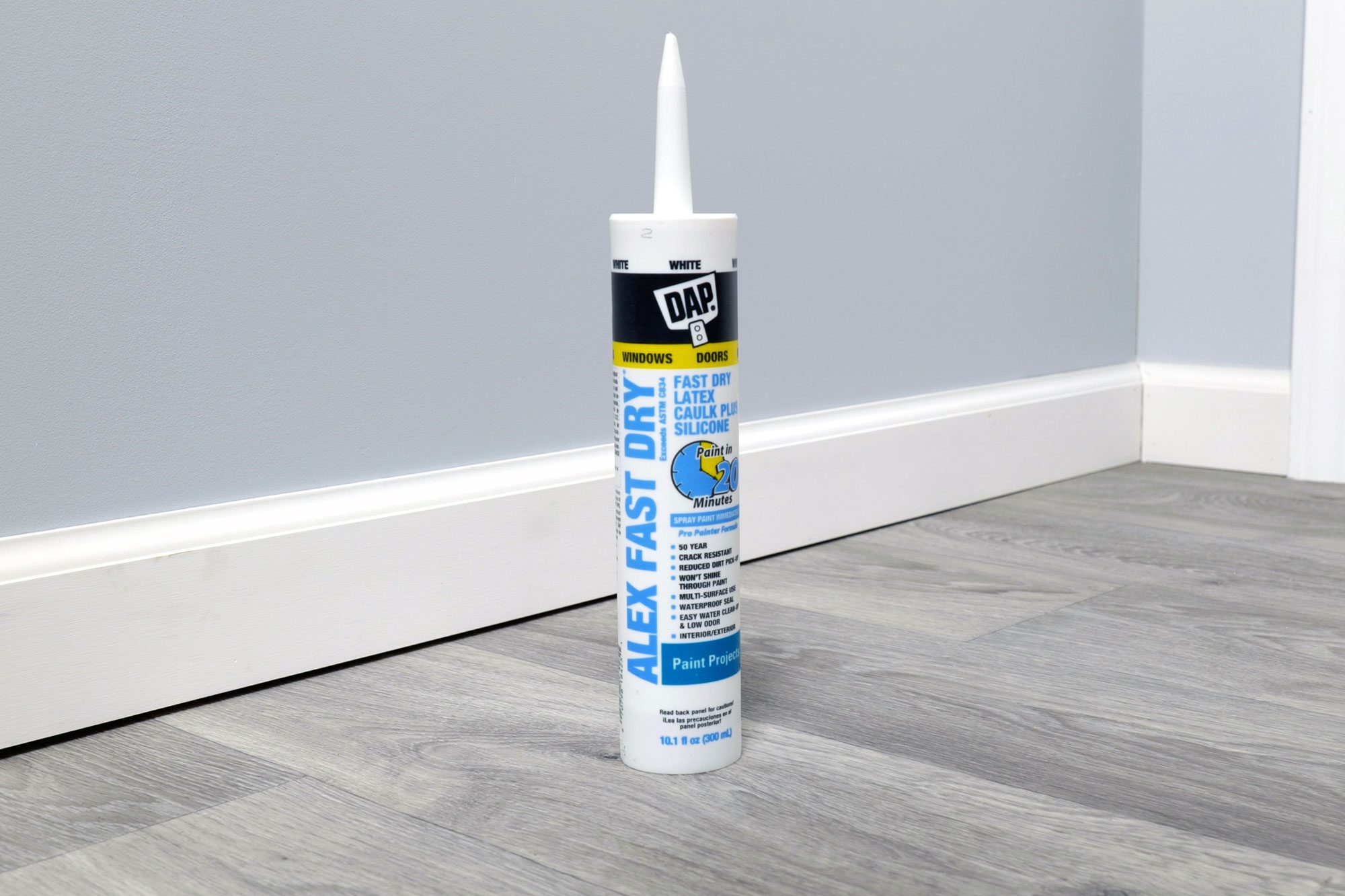 A tube of caulk stands upright on a gray floor, beside a white baseboard, against a light blue wall, indicating preparation for sealing gaps.