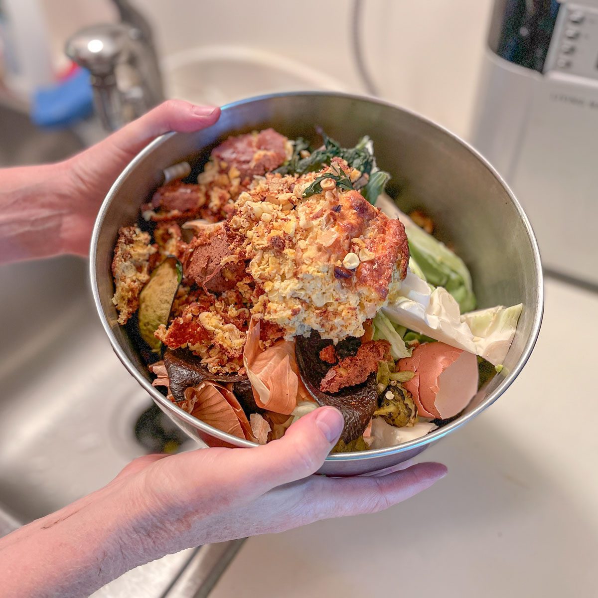 A person holds a bowl filled with food scraps and compost in a kitchen near a sink, preparing to dispose of the waste.