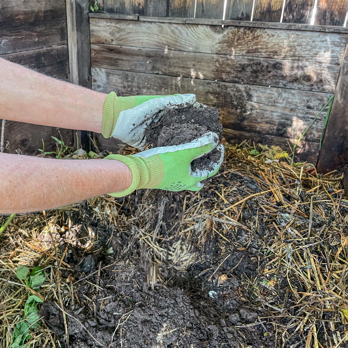 Hands wearing green gloves hold freshly dug soil, preparing to mix it with compost in a natural garden setting surrounded by wooden fencing and straw.