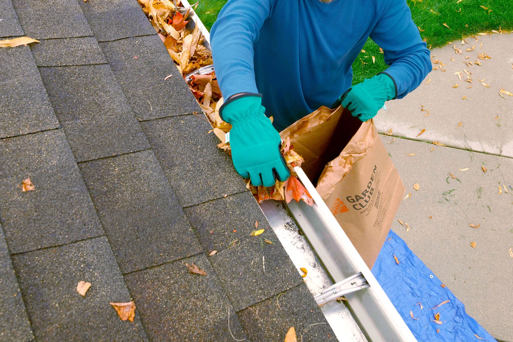 A gloved person is clearing leaves from a gutter into a brown bag while standing on a roof, surrounded by fallen leaves and a blue tarp below.