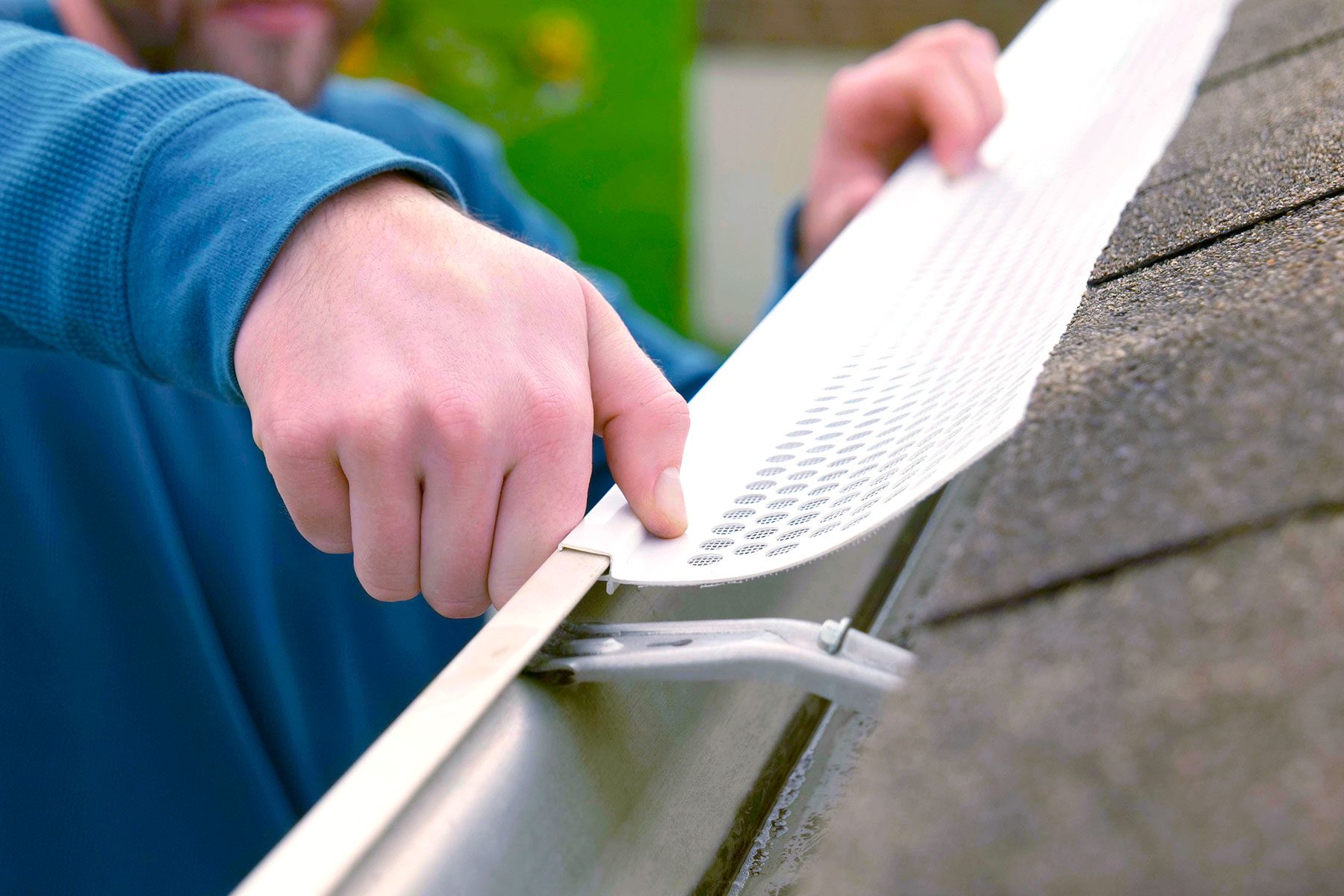A hand is installing a white gutter guard onto a metal gutter, positioned on a roof. The background shows greenery and a clear sky.
