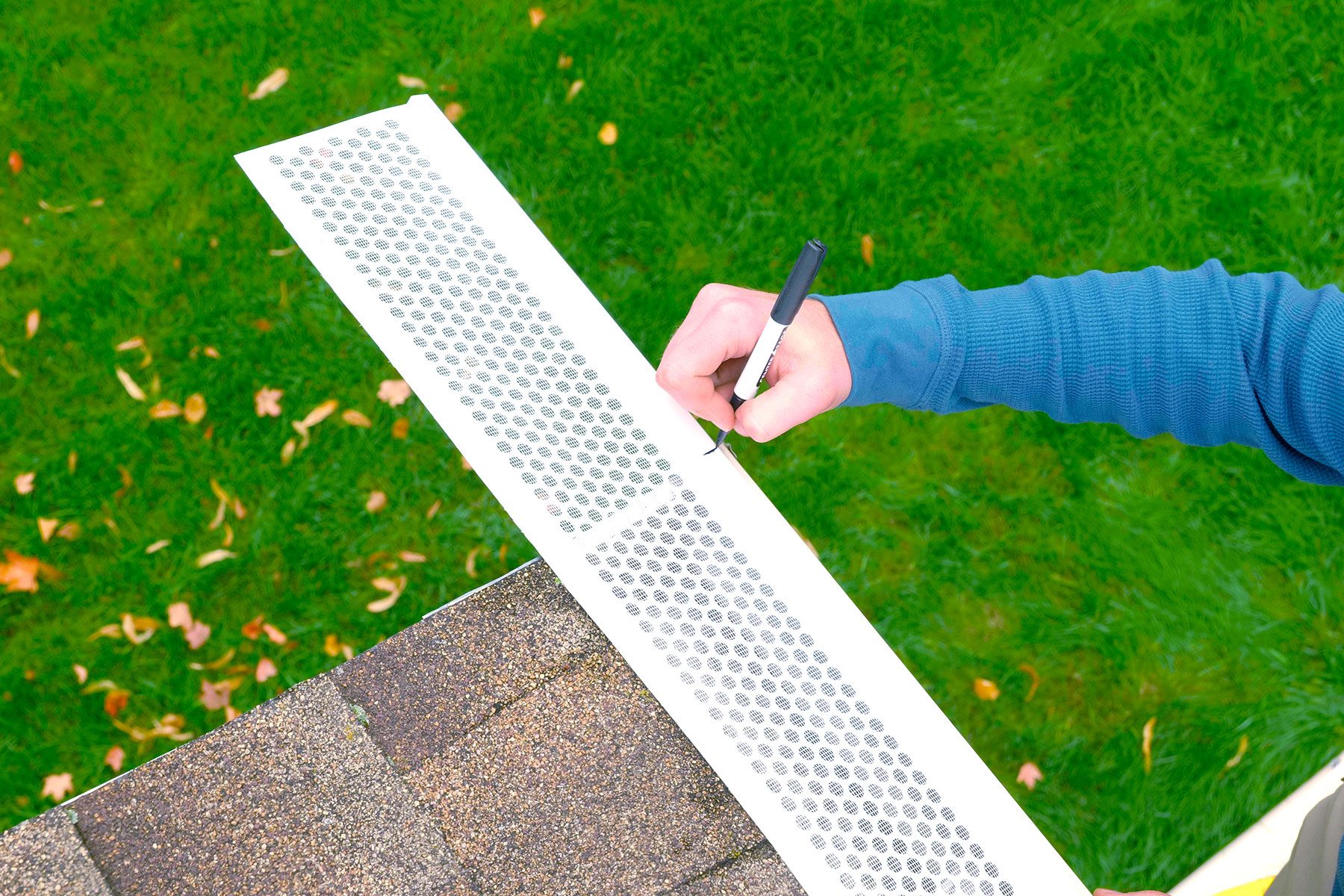 A hand holds a marker, marking measurements on a perforated metal strip situated on a roof's edge, with a grassy yard and fallen leaves below.