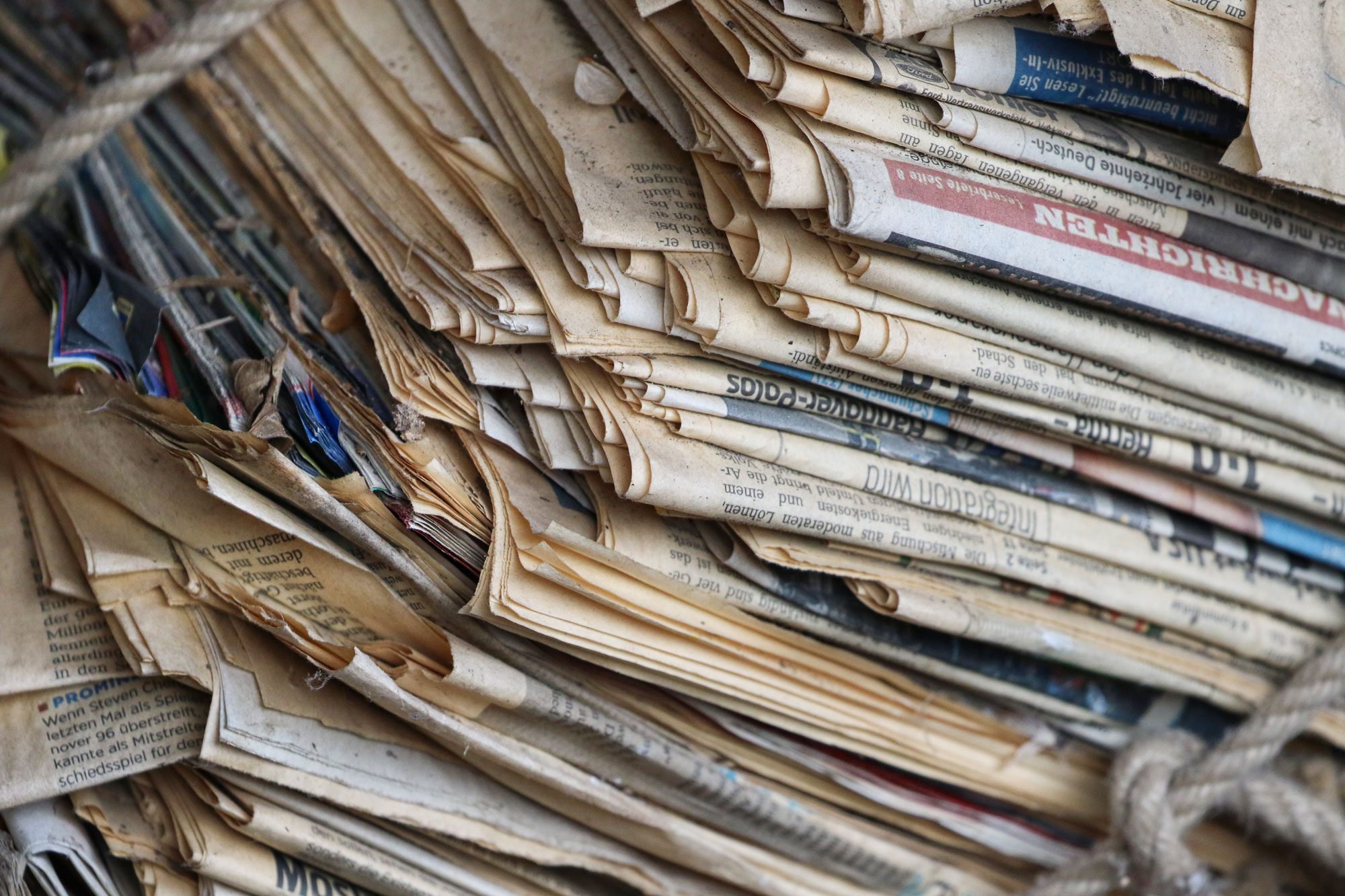 A bundle of aged newspapers is tightly tied with rope, showing yellowed pages and various colors peeking through, resting in a cluttered environment.