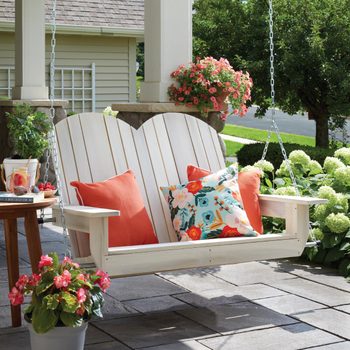 A white wooden swing with colorful cushions gently sways, surrounded by a stone patio, potted flowers, and a lush green landscape.