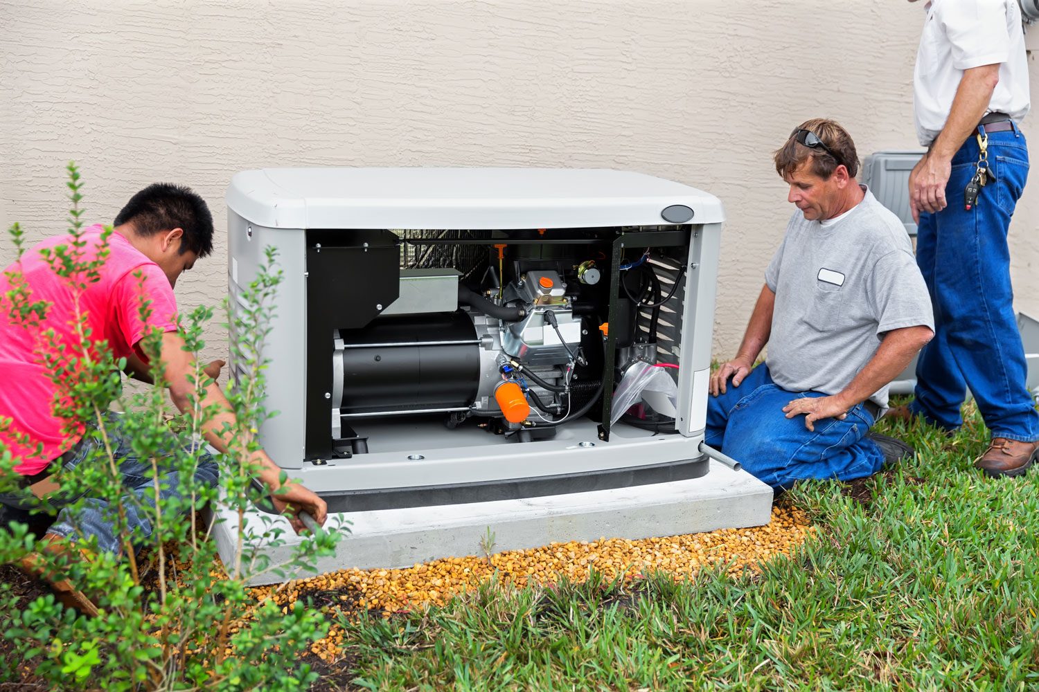 Two technicians are examining an open generator, surrounded by green grass and small rocks, while working near a beige wall.