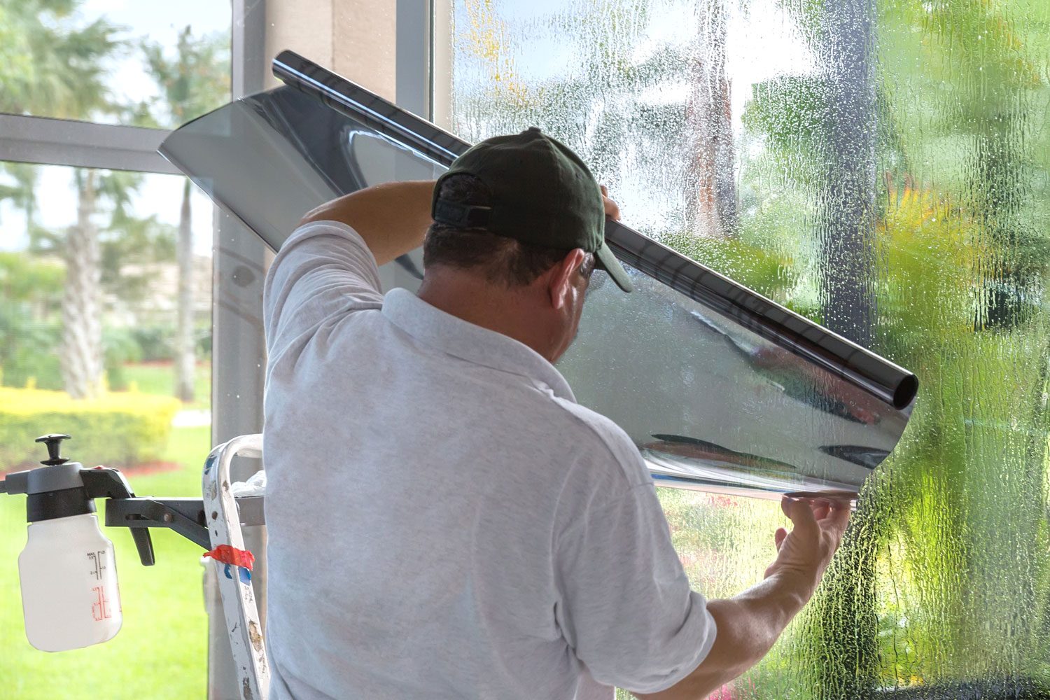 A man applies window film, carefully smoothing it down, in a bright room with tropical plants visible outside through the glass.