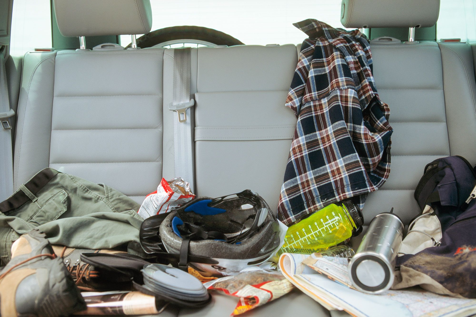 A cluttered backseat features scattered clothes, shoes, a helmet, snacks, and a water bottle, suggesting recent outdoor activities or travel.
