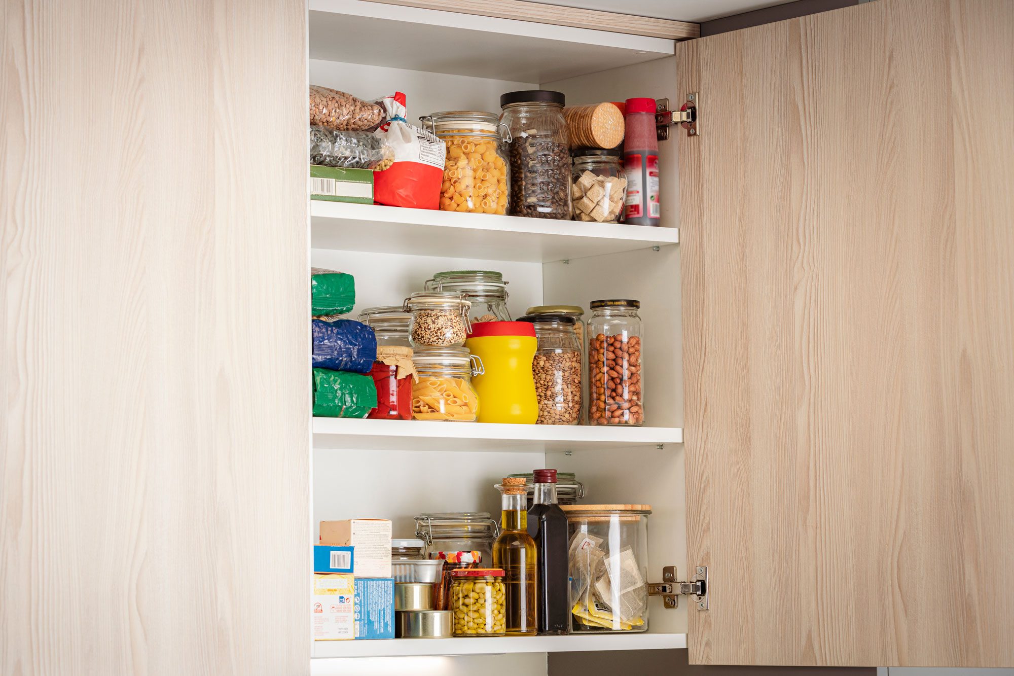 A kitchen cabinet holds various containers and jars filled with grains, pasta, spices, and condiments, organized on white shelves against a light wood background.