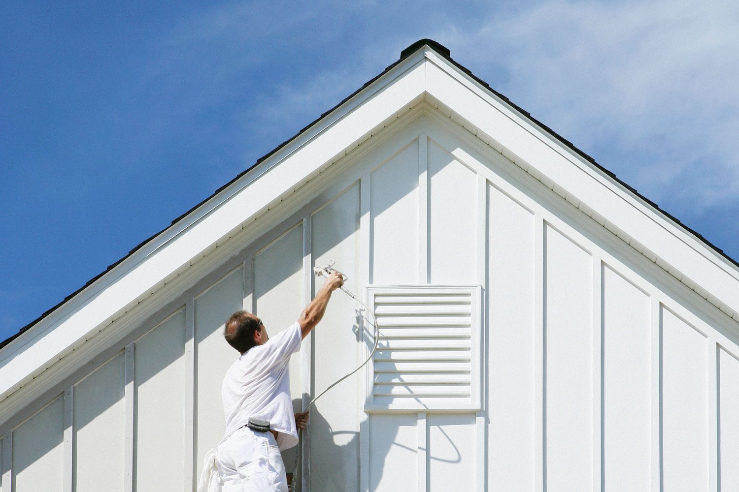 A man paints the side of a white house, using a sprayer, under a clear blue sky with few clouds.