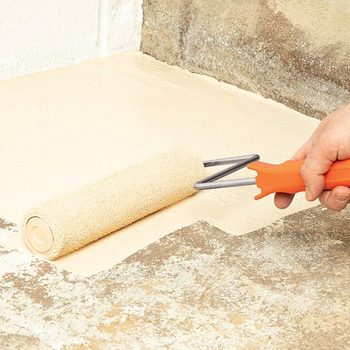 A hand holds a paint roller, applying light beige paint to a concrete surface, which shows previous faded colors and textural variations.