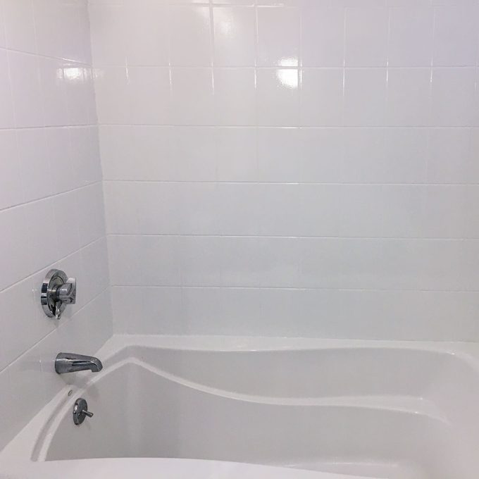 A white bathtub is positioned against tiled walls, with a faucet and controls visible. Light reflects off the smooth surfaces, creating a clean, minimalist look.