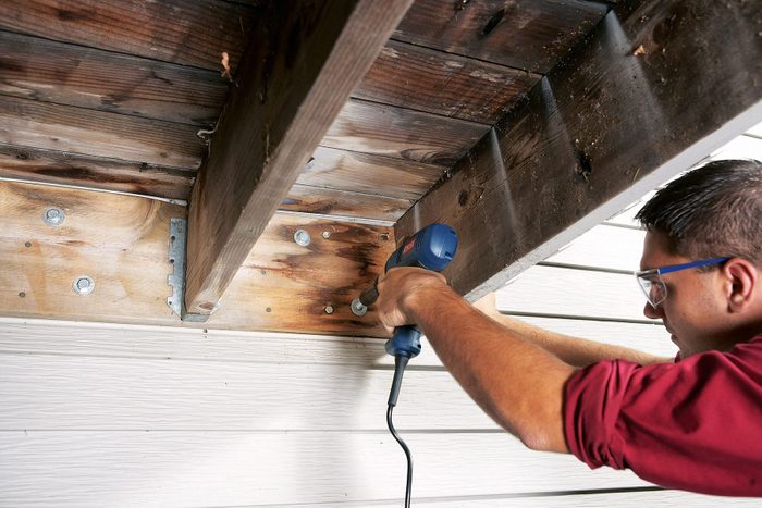 A person in a red long-sleeve shirt is using a power drill to secure bolts into wooden beams on a deck or patio structure. The scene is viewed from below, showing the detailed construction and alignment of the wooden planks and bolts.