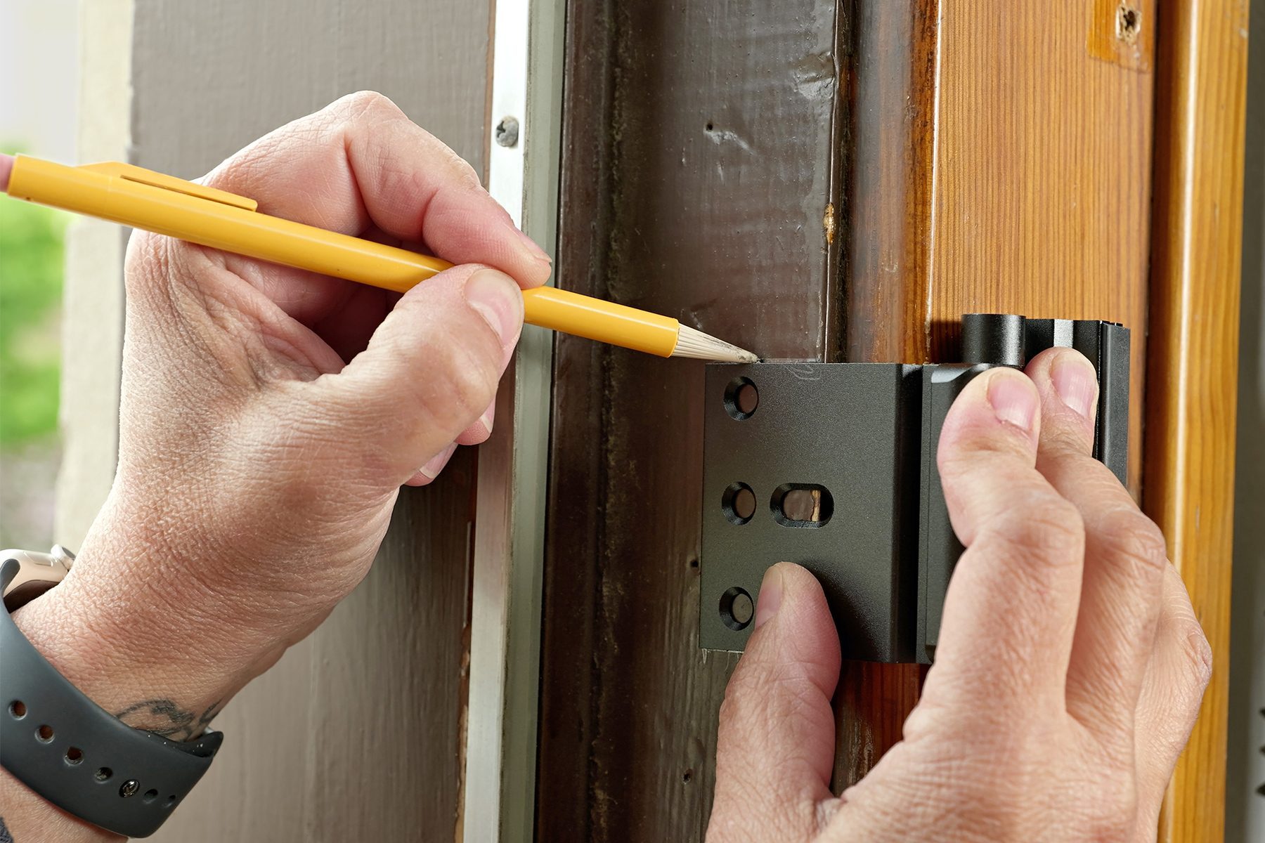 A person is using a pencil to mark the placement of a door hinge on a wooden door frame. One hand holds the hinge in place while the other hand marks the screw holes with the pencil. The edge of the open door is visible on the right.