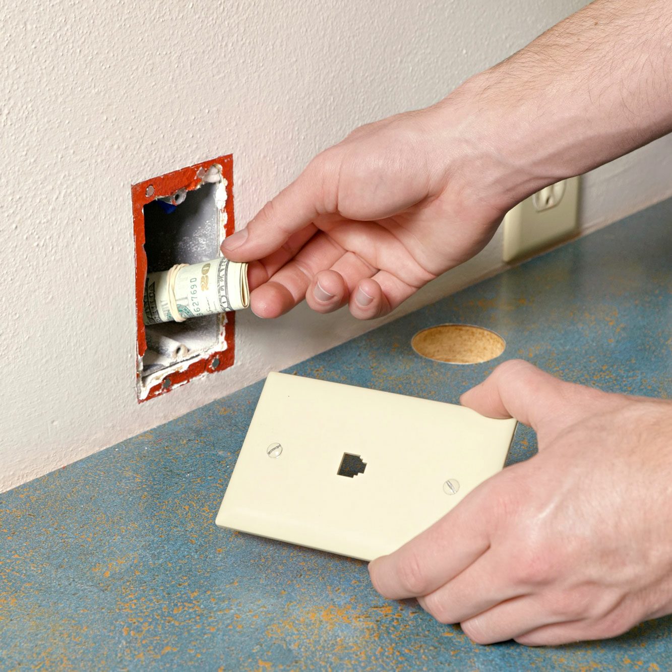 a person's one hand is holding a white electrical socket cover, while the other hand is inserting a rolled-up banknote into an open electrical outlet box in the wall; The outlet box has exposed wires and no socket installed; Nearby, there is a green plant and a white patterned vase on the countertop;