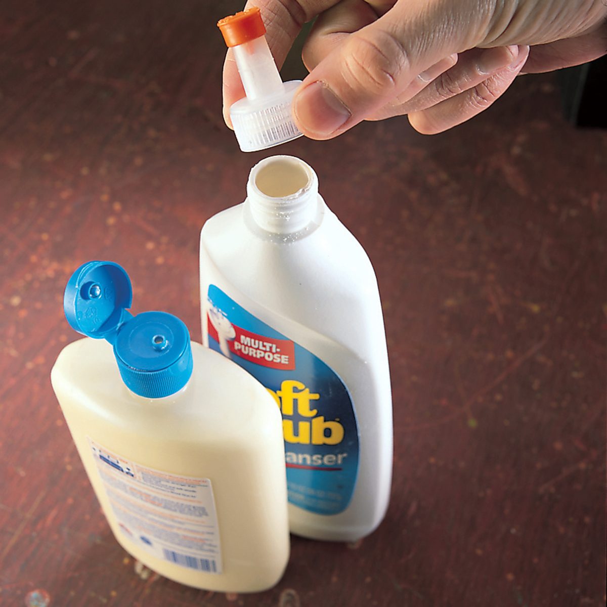 A person is holding the cap of an open plastic bottle labeled "Soft Scrub Cleanser" next to another plastic bottle with a blue cap. Both bottles are on a dark surface. The focus is on the bottles and the caps.