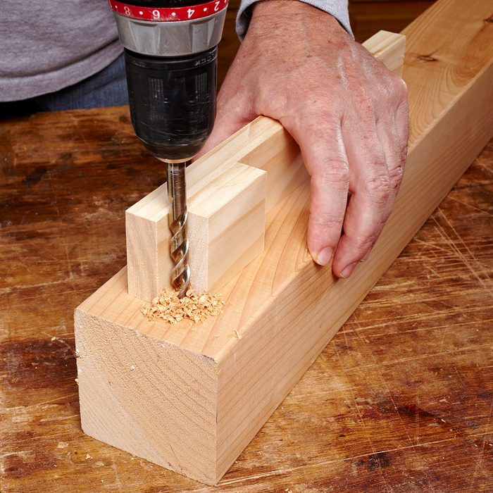 A person using a drill to create a hole in a piece of wood, clamped onto a wooden workbench. Sawdust is visible around the drill bit as it penetrates the rectangular wooden block. The person's hands are steadying the wood.