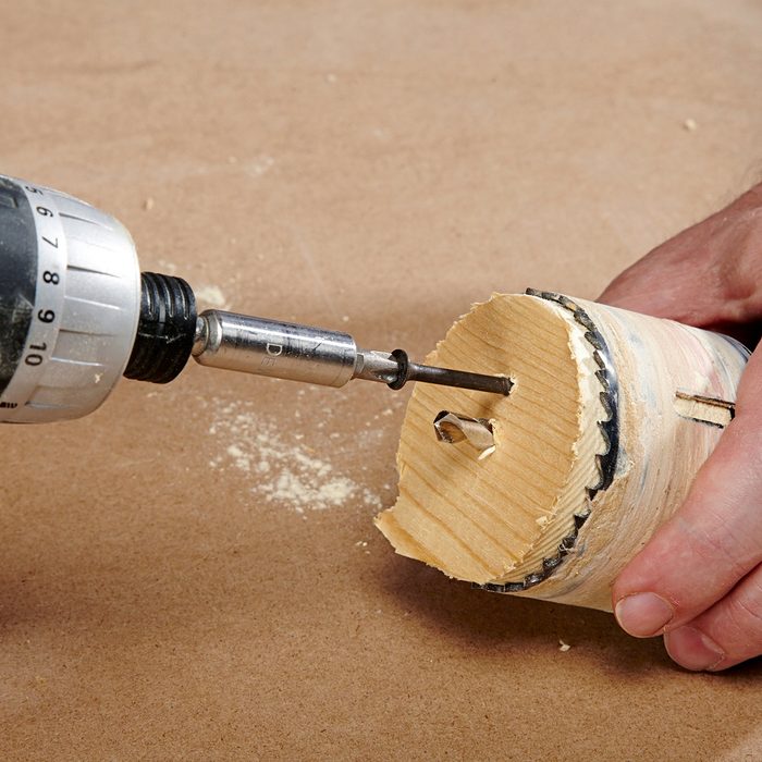 A person's hand holding a piece of wood as a drill makes a hole in it. The wood is cylindrical with a cut groove, and wood shavings are visible. The drill bit is in motion against a brown surface.
